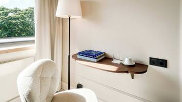 Une chambre d’hôtel avec une fenêtre, une chaise, une table de chevet avec un livre et un verre, et une lampe.