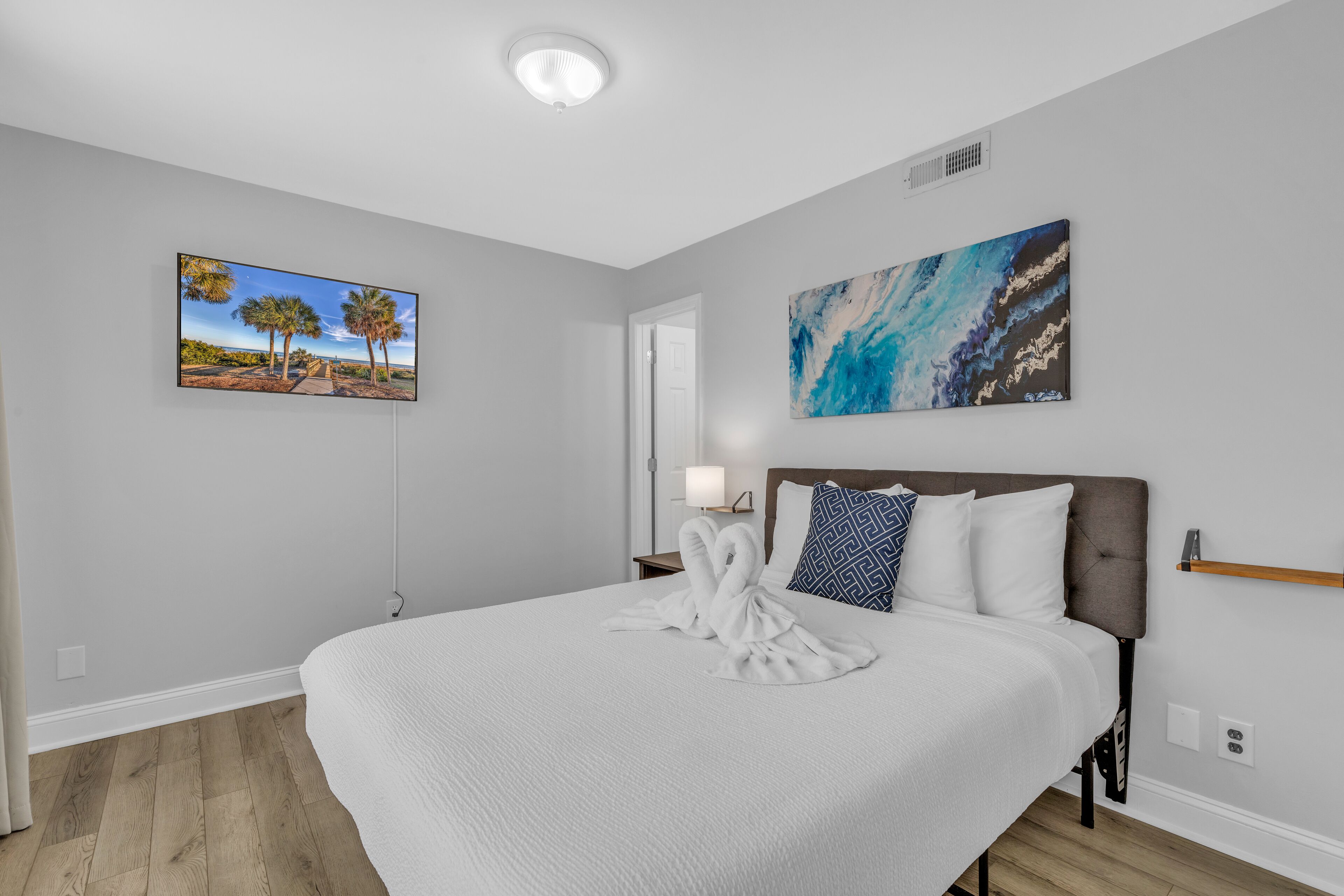 A hotel room with a large bed, a painting of a beach scene, a painting of a blue abstract design, and a white towel on the bed.