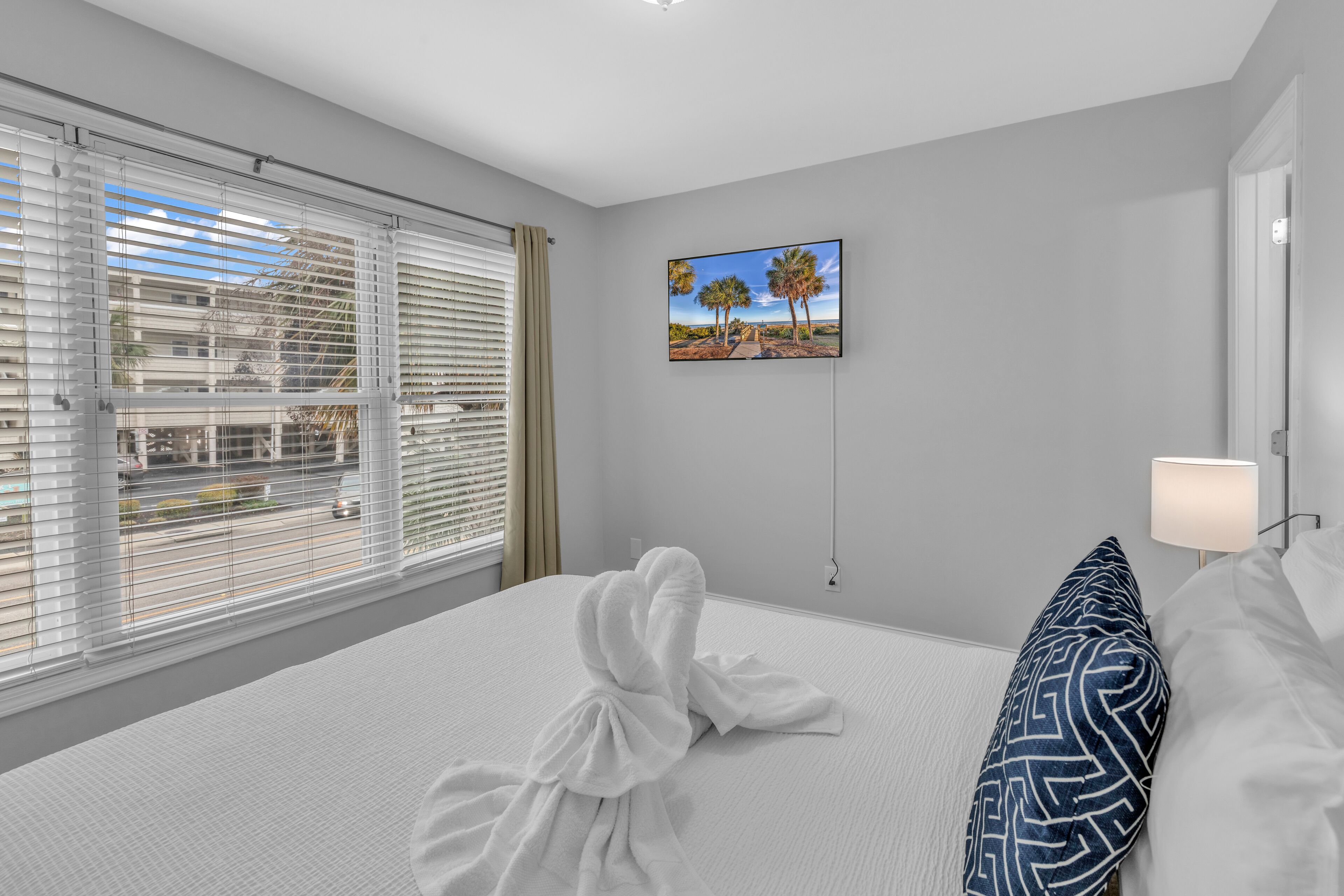 A hotel room with a large bed, a painting of palm trees, and a view through the window.