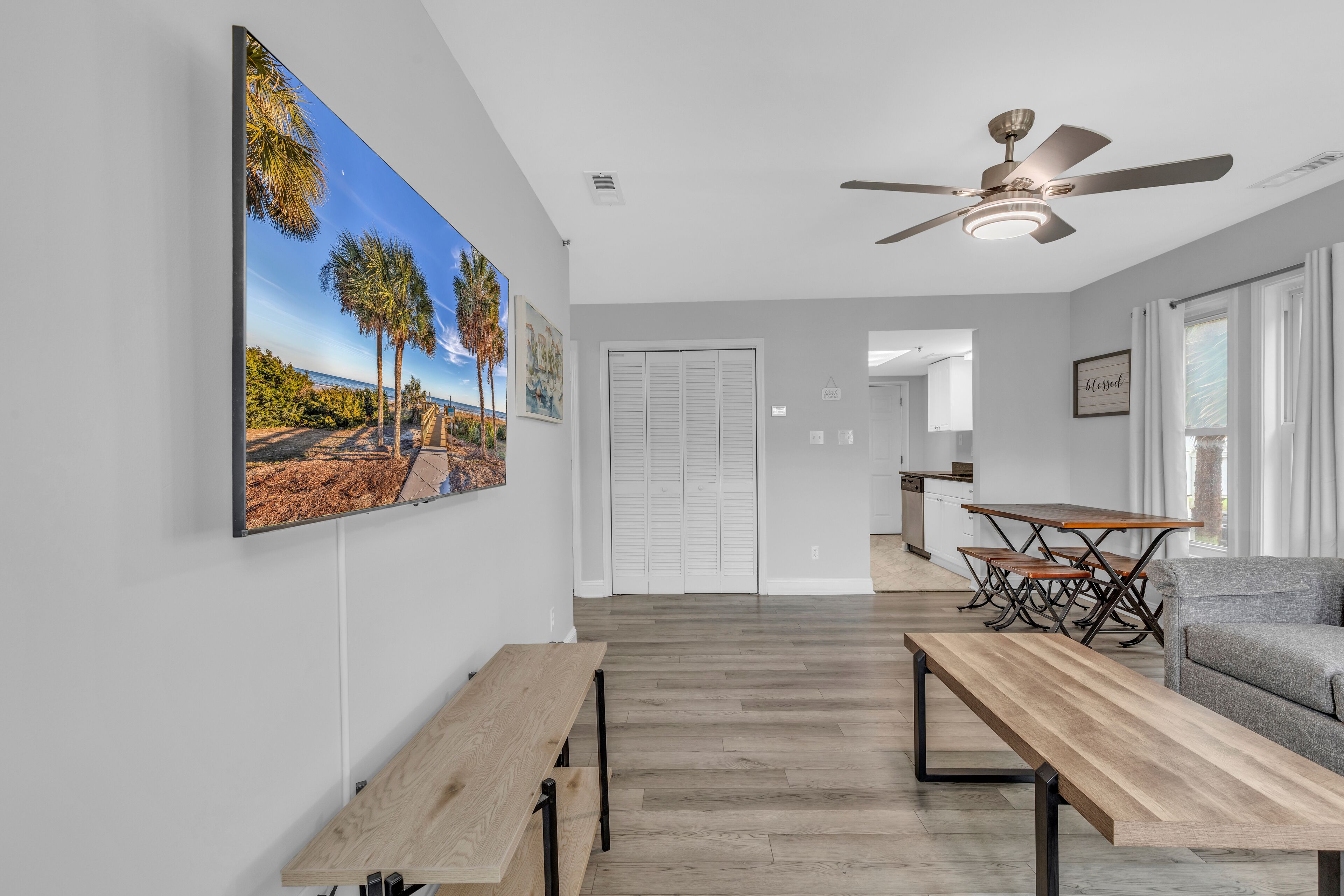 A modern living room with a wooden bench, a dining table, and a gray sofa. There is a ceiling fan, a large painting of a beach scene, and a view into the kitchen area.
