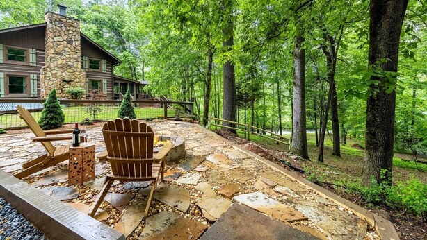 Un patio de piedra con una silla Adirondack de madera y una botella de vino, rodeado de un área boscosa y un edificio de piedra al fondo.