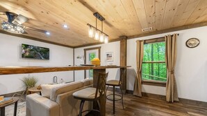 Interior acogedor de una cabaña con techo de madera, una zona de comedor con banco y sillas, una encimera de cocina y un amplio ventanal con cortinas.