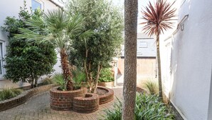 A courtyard with a brick pathway, surrounded by various plants and trees, including palm trees and a large ficus. - 11 The Craighurst (Reydon)
