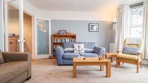A living room with a sofa, armchair, coffee table, bookshelf, and a door leading to another room. - 11 The Craighurst (Reydon)