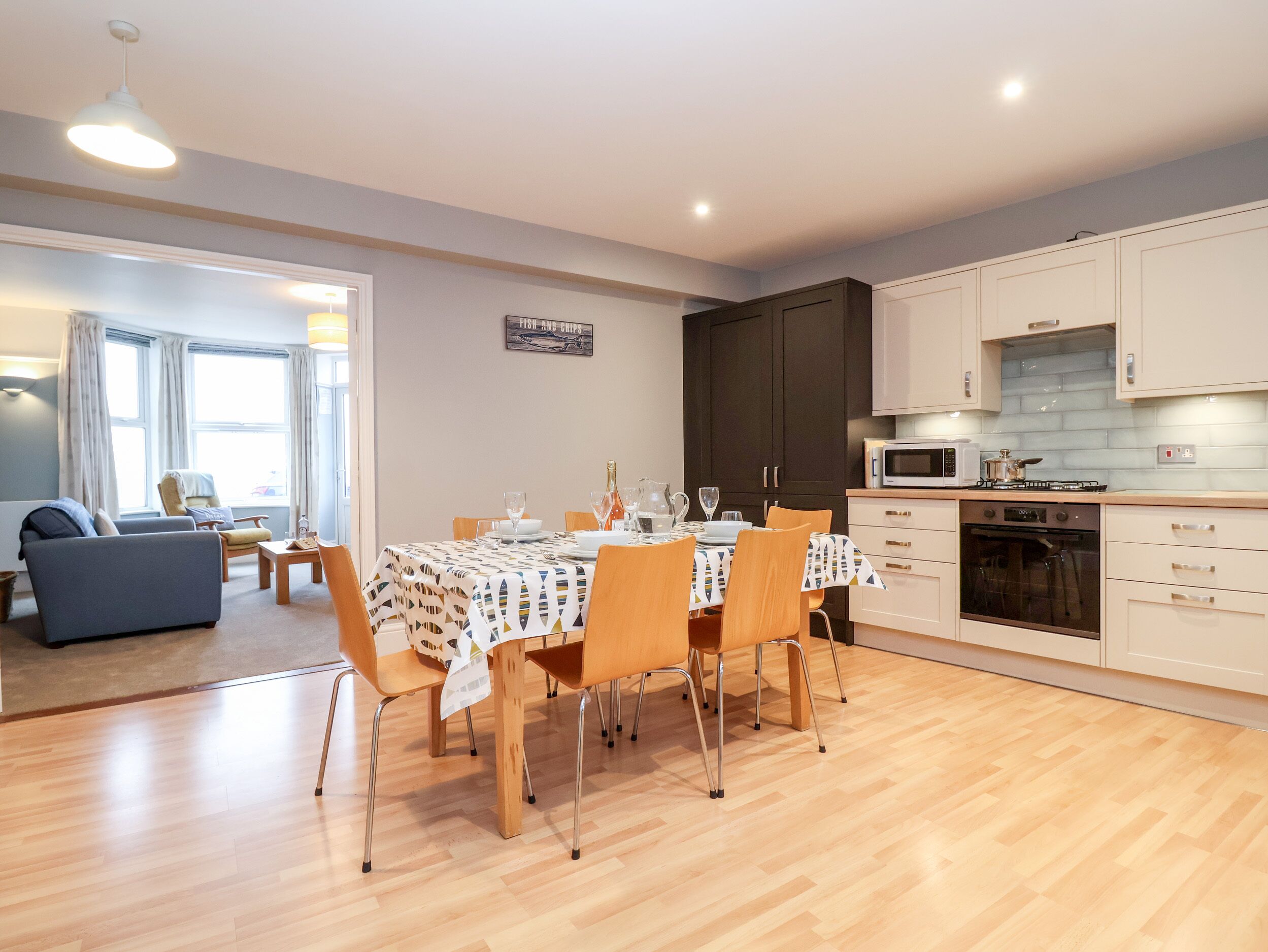 A modern kitchen with wooden cabinets, a dining table set for four, and a view into a living area with a sofa and a window.