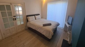 Une chambre à coucher comprenant un lit, un téléviseur, une fenêtre avec des stores et une porte dotée de panneaux en verre dépoli.