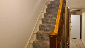 Un escalier dont les marches sont recouvertes de moquette et qui possède une rampe en bois, menant à une porte. On aperçoit un réfrigérateur blanc à l’arrière-plan.