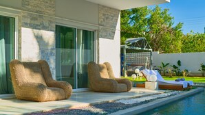A modern outdoor pool area with two wicker lounge chairs, a sunbed, and a glass door leading to a covered patio. - Luxury Villa with Pool,BBQ and Garden Vouliagmeni
 (Voula)