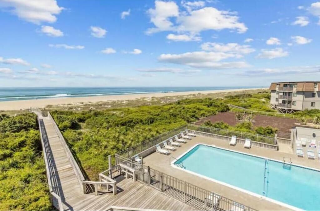A pool area with a view of the beach and ocean, surrounded by greenery and a wooden deck.