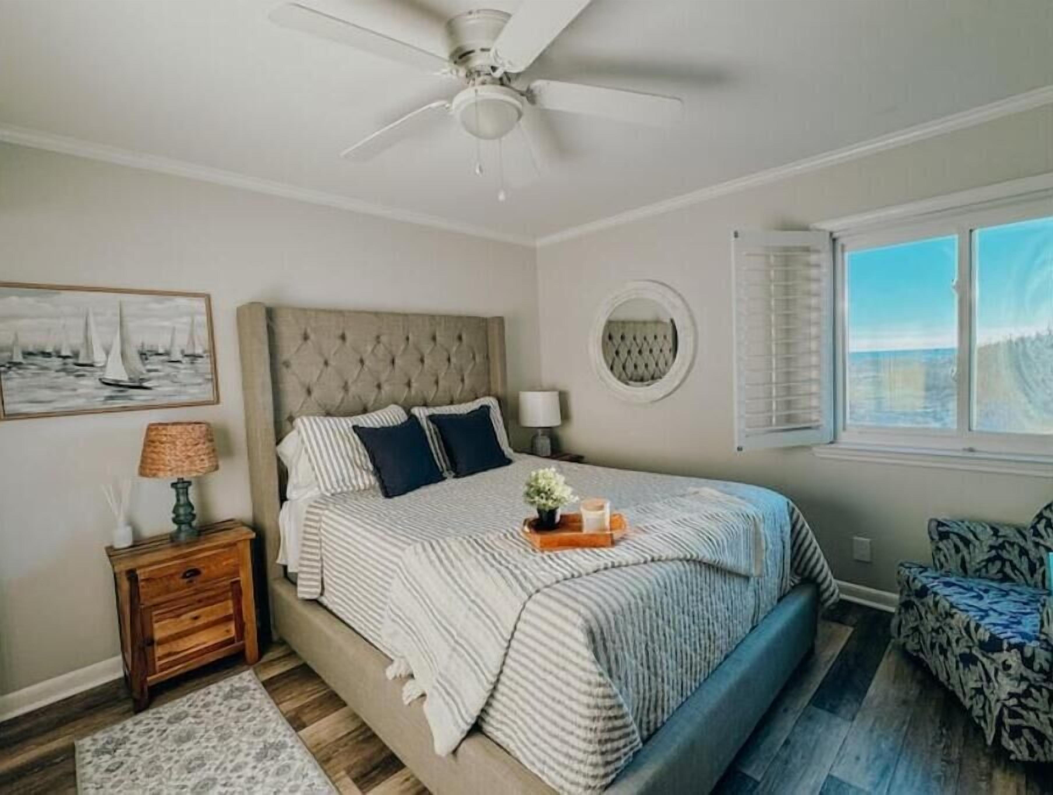 A bedroom with a tufted headboard, a bed with striped bedding, a nightstand with a lamp, a round mirror, a window with blinds, and a patterned chair.
