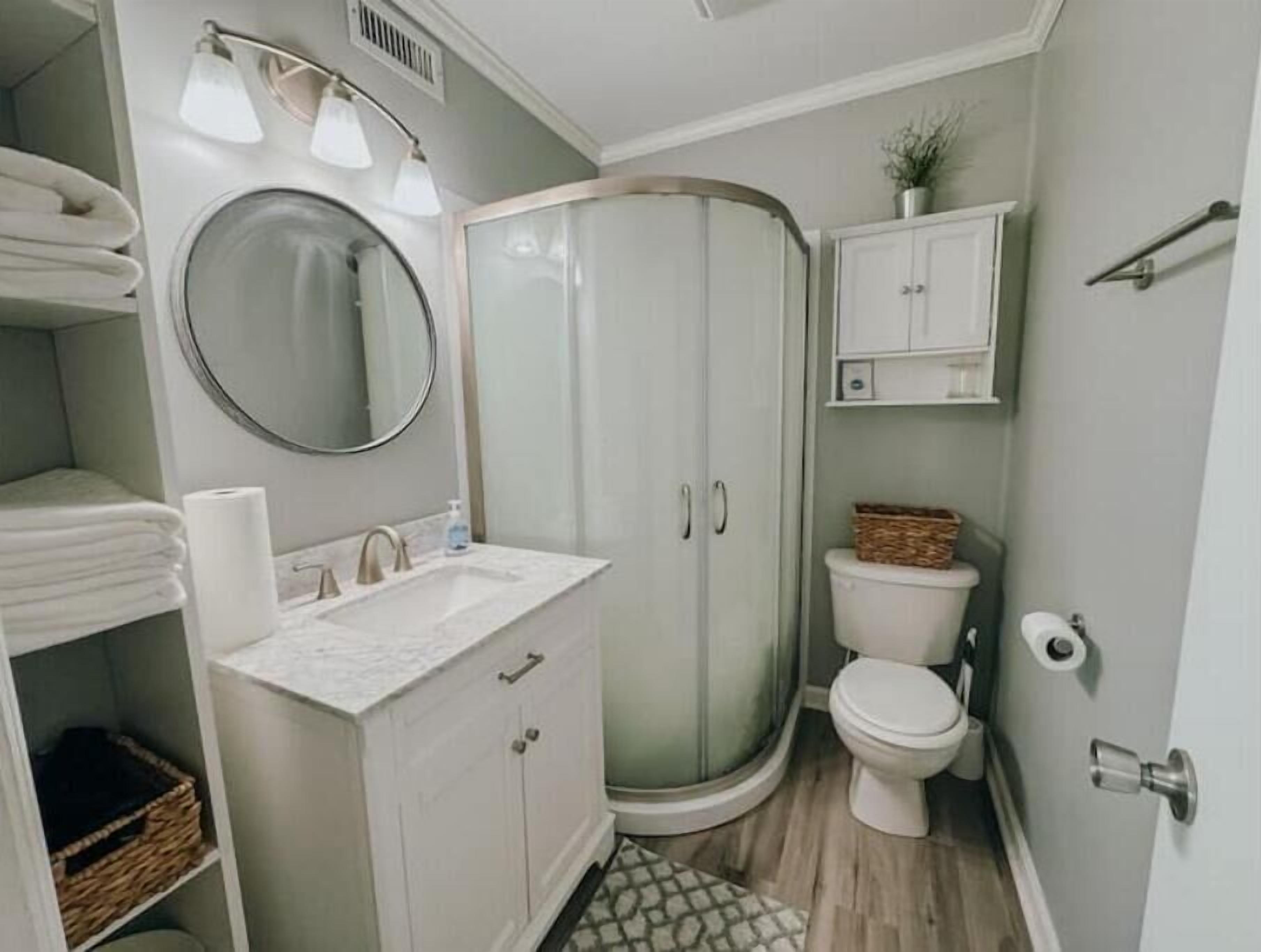A bathroom with a white vanity, a round mirror, a glass-enclosed shower, a toilet, and a small cabinet with toiletries.