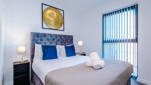 A modern hotel room with a large bed, two nightstands, a framed artwork above the bed, and a window with blinds.