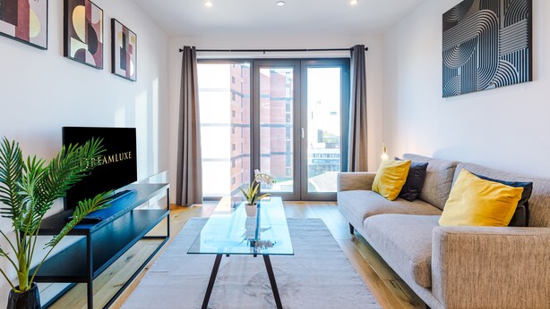 A modern living room with a flat-screen TV on a black stand, a glass coffee table, a beige sofa with yellow cushions, and a large window with curtains.