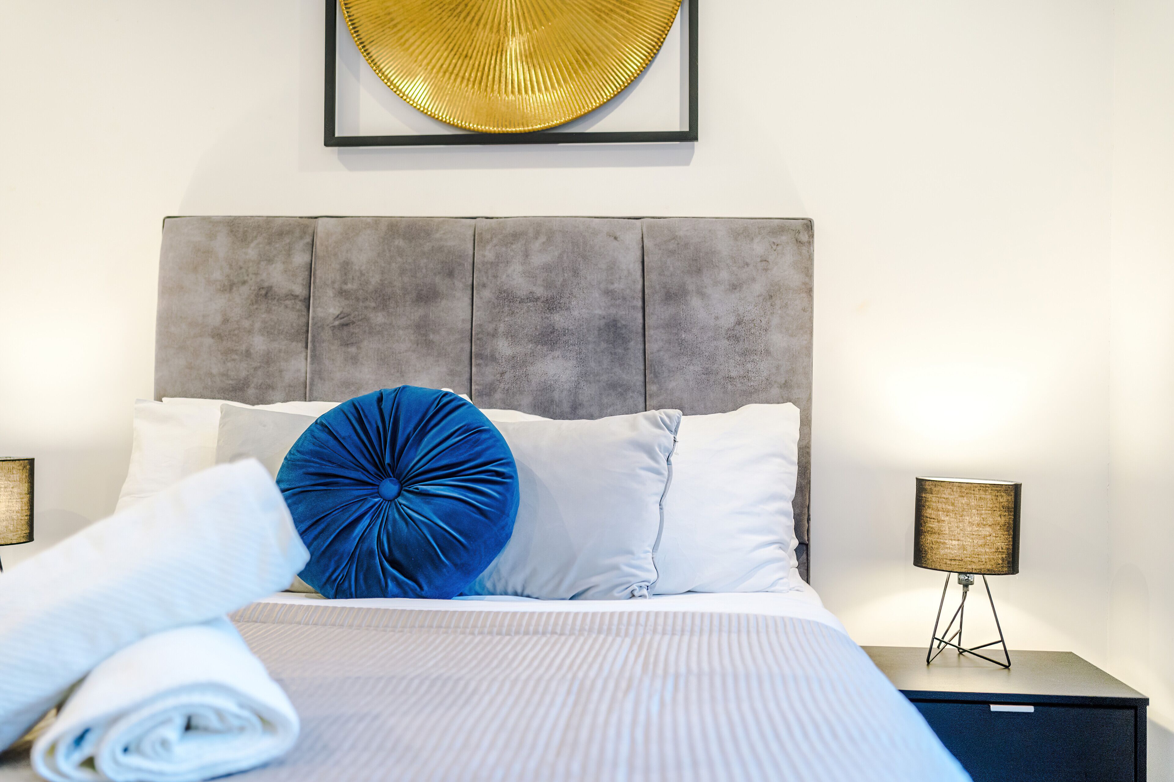 Un lit bien fait, agrémenté d’un coussin bleu, avec un tableau encadré au-dessus et une lampe de chevet posée sur une table de nuit.