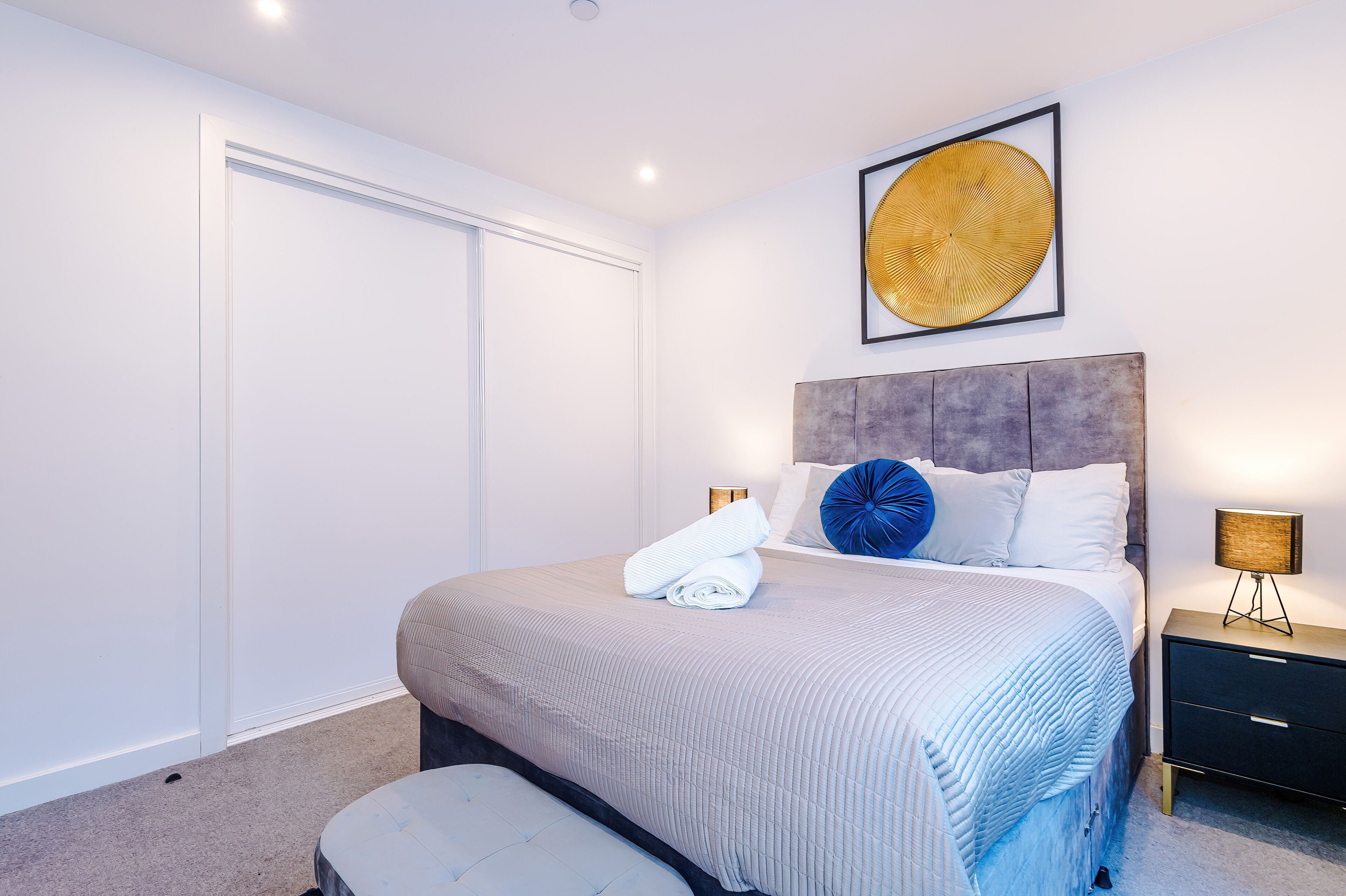 A neatly made bed with a headboard, a bedside table with a lamp, and a large framed artwork on the wall.