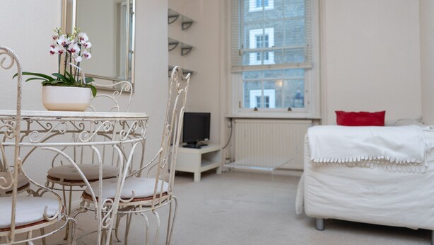 A room with a white bed, a white chair, a white table with a potted orchid, a large mirror, a window with blinds, and a red pillow. - Beautiful 1-bed Apartment in London (London)