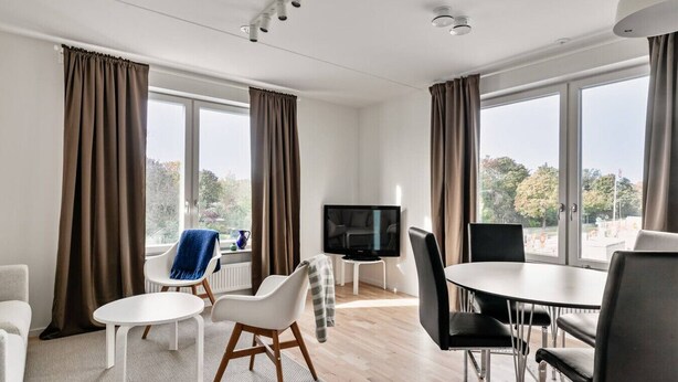 A modern hotel room with a flat-screen TV, a dining table, two chairs, a sofa, and large windows with curtains. - Stylish Urban Home in Stockholm (Stockholms län)