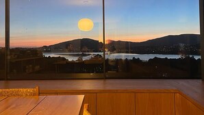 A room with a large window offering a view of a sunset over a body of water and hills.