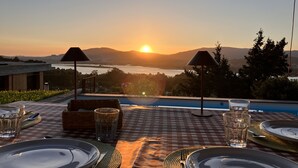 A dining table set for two with a view of the sunset over a body of water and mountains.