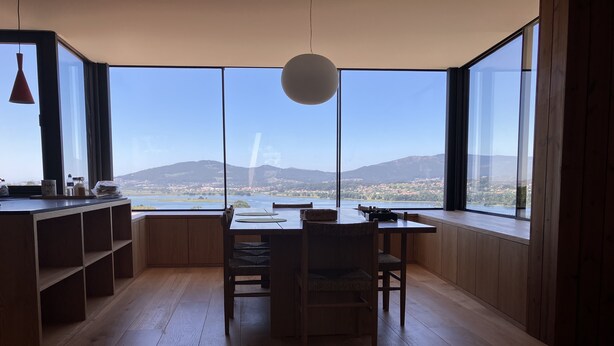 A room with a wooden floor, a dining table with chairs, a large window offering a view of a lake and mountains, and a pendant light.