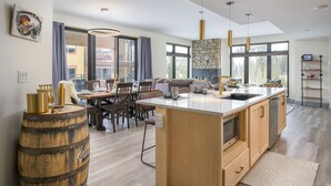 A modern kitchen with a large island, wooden cabinets, and a stone backsplash. There is a dining area with a table and chairs, and large windows providing natural light.
