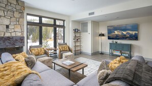 A spacious living room with a stone fireplace, a large window overlooking a snowy landscape, a comfortable sofa set, and a mounted painting.