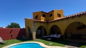 A swimming pool with a stone deck, a yellow building with arches, and a clear blue sky. - Casa Lares (Casas de Don Pedro)