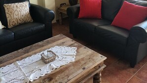 A living room with two black sofas, one with a patterned cushion and the other with red cushions, a wooden coffee table with a lace table runner, and a lamp on a side table. - Casa Lares (Casas de Don Pedro)