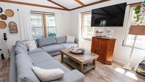 A modern living room with a grey sectional sofa, a wooden coffee table, and a large TV mounted on the wall. - Ocean View, Semi-Oceanfront, Screened In Porch, Beach Access (Avon)