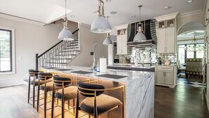 A modern kitchen with a marble island, bar stools, pendant lighting, and a staircase.