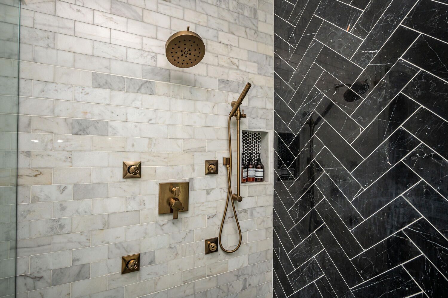 A bathroom with a white marble tiled wall, a black marble tiled wall, and a shower with a gold showerhead and handheld shower.