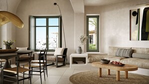 A spacious living area with a high ceiling, a dining table with chairs, a sofa, a coffee table, and a large window offering a view of a coastal town. - Patina & Vivera Estate Santorini (Pyrgos)