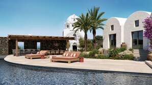 A poolside area with lounge chairs and a pergola, a white building with a dome, and a palm tree. - Patina & Vivera Estate Santorini (Pyrgos)