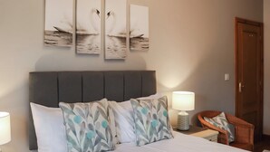 A neatly made bed with white linens, two decorative pillows with a leaf pattern, a grey upholstered headboard, a bedside lamp, and a wooden chair with a cushion.
