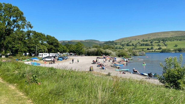 A lakeside campsite with tents and canopies, surrounded by greenery and hills.