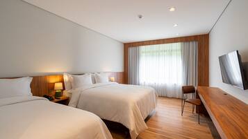 Quarto de hotel com duas camas, uma escrivaninha de madeira, televisão e uma janela grande com cortinas.