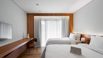 Une chambre d’hôtel moderne avec un bureau en bois, deux lits, une télévision et une grande fenêtre agrémentée de rideaux légers.
