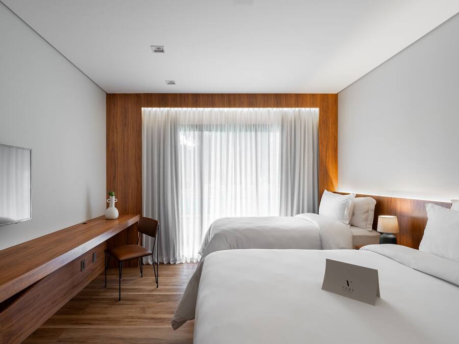 Une chambre d’hôtel moderne avec un bureau en bois, deux lits, une télévision et une grande fenêtre agrémentée de rideaux légers.