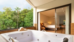 A modern hotel room with a large bathtub, a balcony with a view of lush greenery, and a sliding glass door leading to a bedroom. - Vert Hotel Spa (Itatiaia)