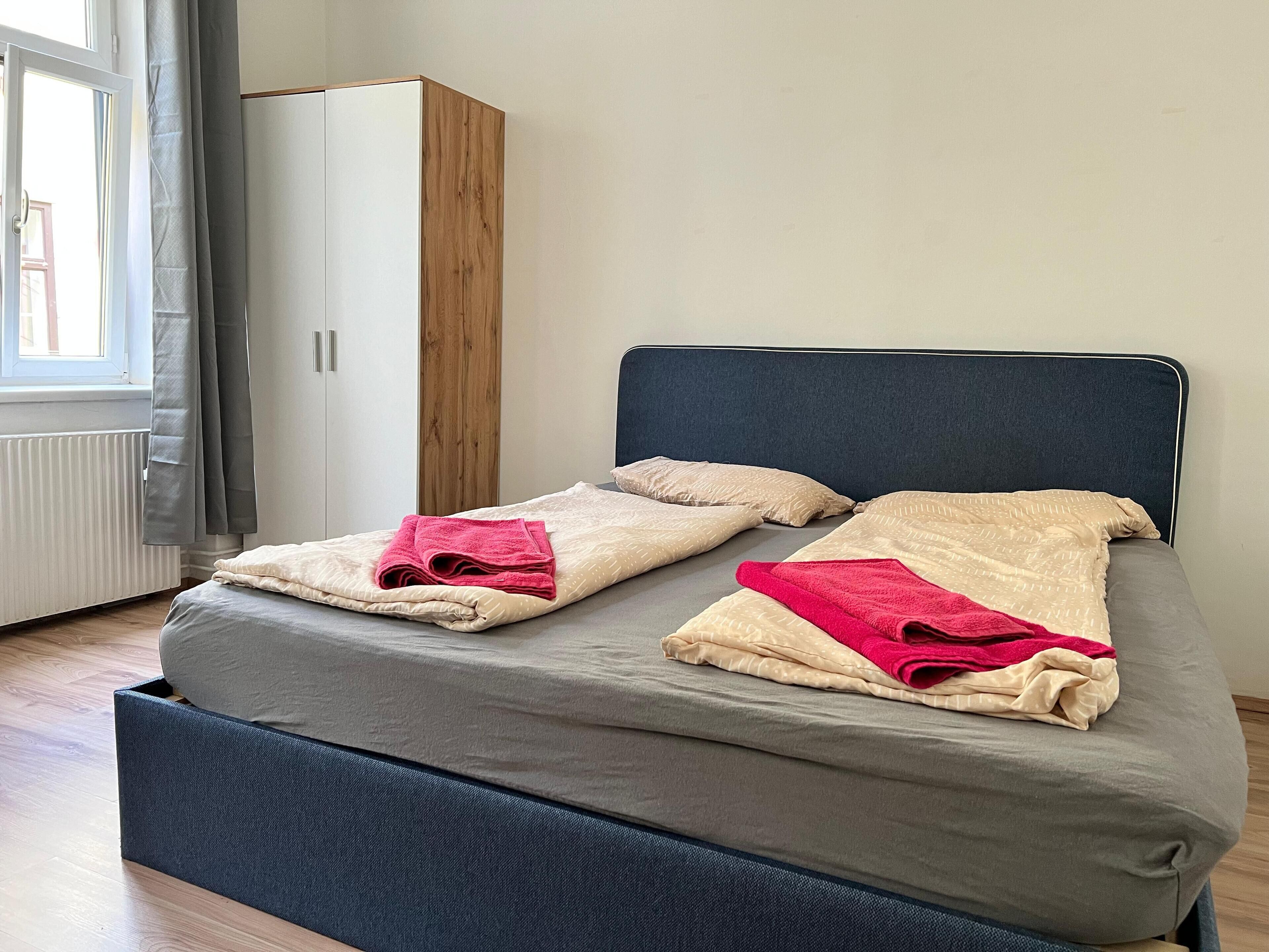 Un lit bien fait, avec deux oreillers et deux serviettes pliées, une tête de lit sombre et une armoire agrémentée d’un élément décoratif en bois.