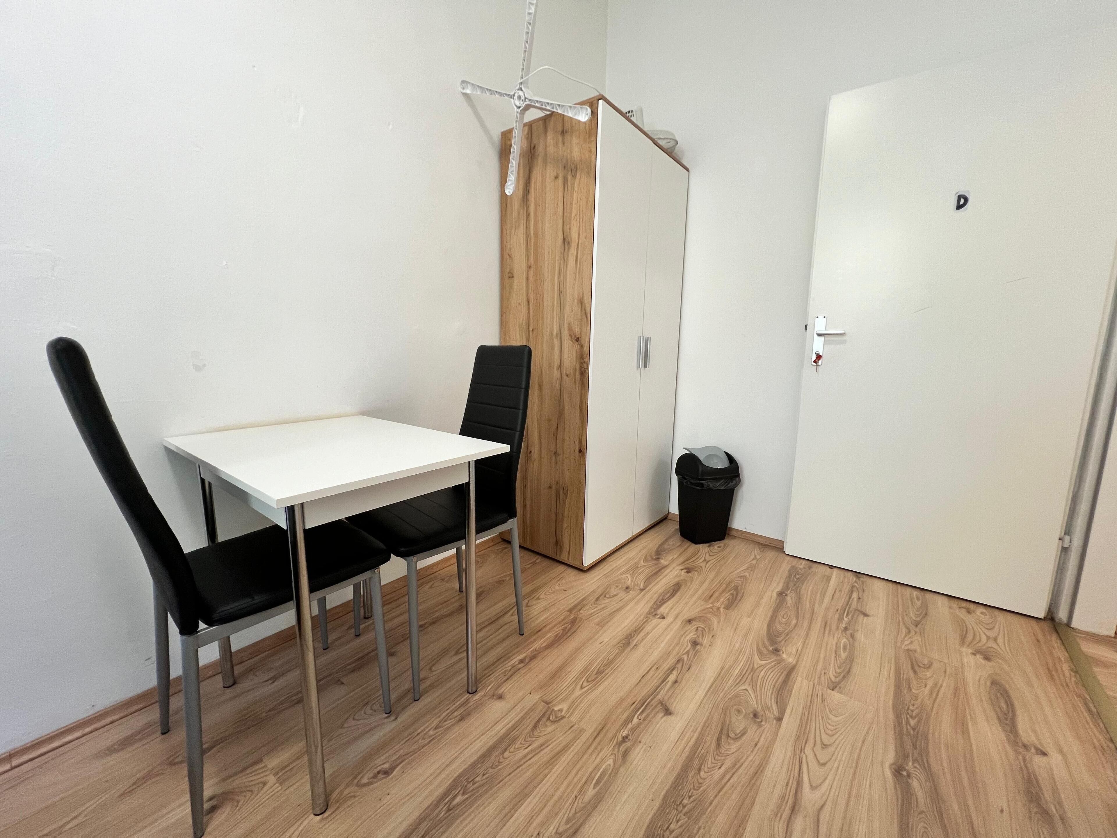 Una stanza piccola con un tavolo bianco e due sedie nere, un armadio in legno, un cestino nero e una porta bianca.