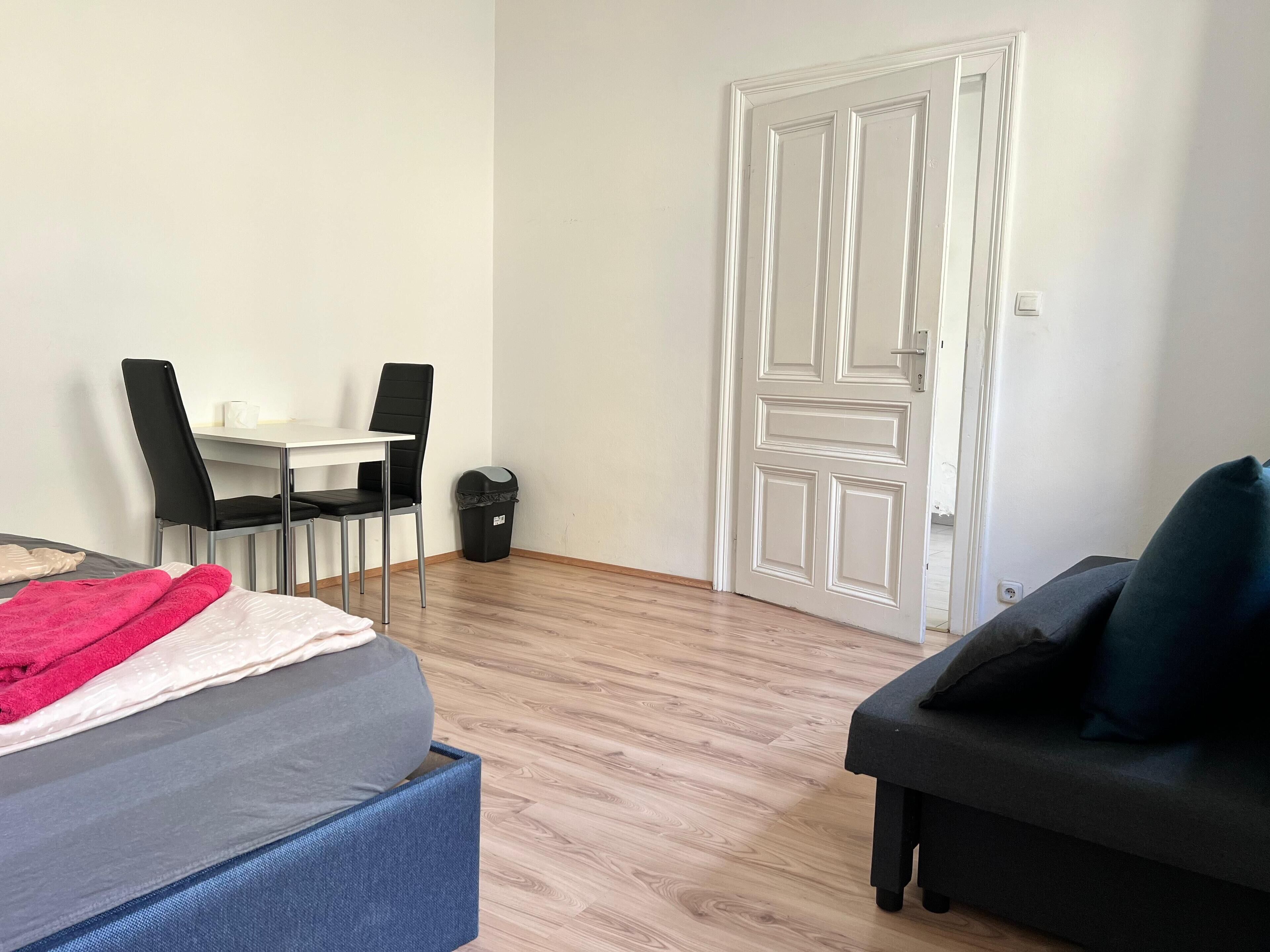 Una habitación con cama, un escritorio con dos sillas, un cubo de basura negro y una puerta.