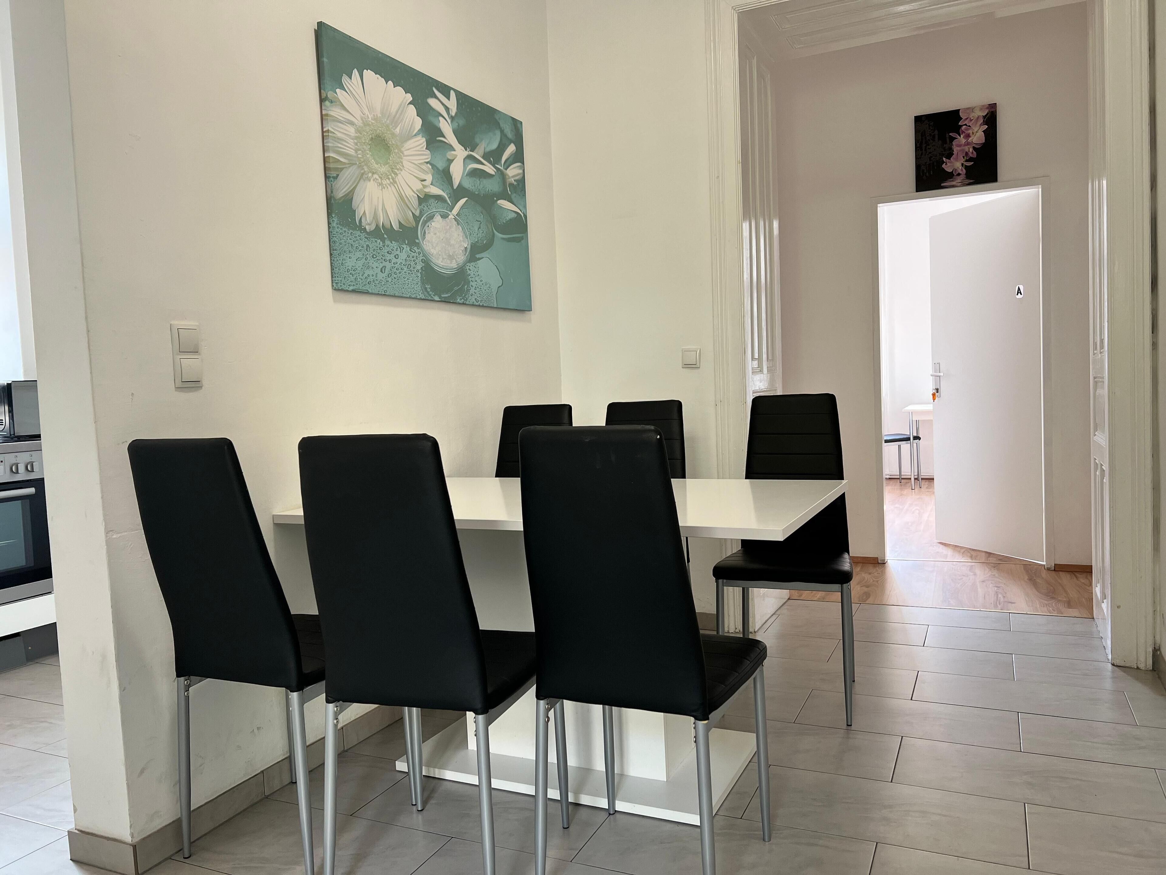 A dining area with a white table and four black chairs, a painting of flowers on the wall, and a kitchen visible in the background.