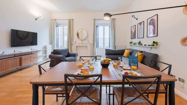 A modern living room with a dining table set for breakfast, a fireplace, and a TV mounted on the wall.
