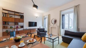 A modern living room with a sofa, a dining table set with breakfast, a bookshelf, a flat-screen TV, and a fireplace.