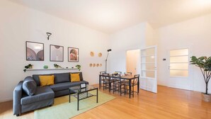 A modern living room with a grey sofa, a coffee table, and a dining area with a wooden table and chairs. There are framed artworks on the wall, a potted plant, and a door leading to another room.