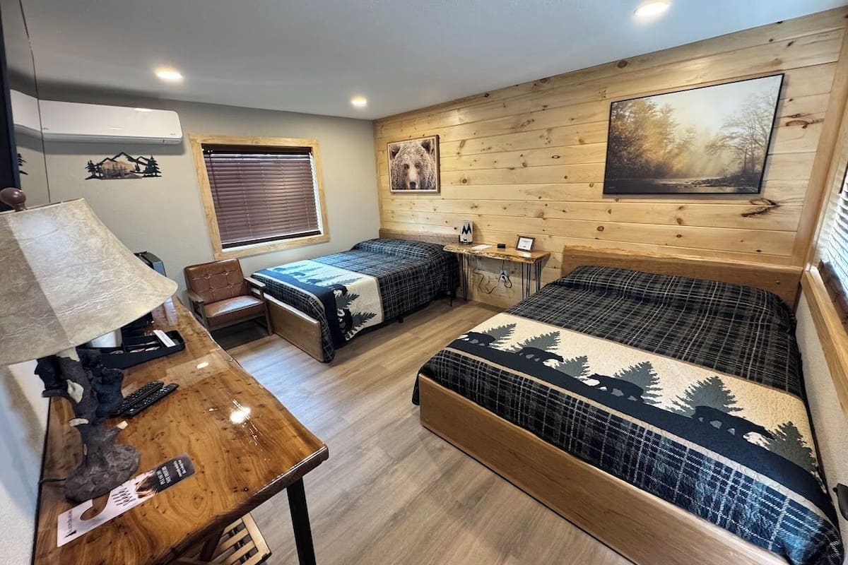 A room with two beds, a wooden desk, a lamp, and a window with blinds.