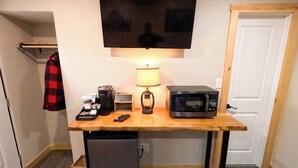 A compact kitchenette with a microwave, coffee maker, and a small refrigerator. A television is mounted above the counter, and a lamp is placed on the table.