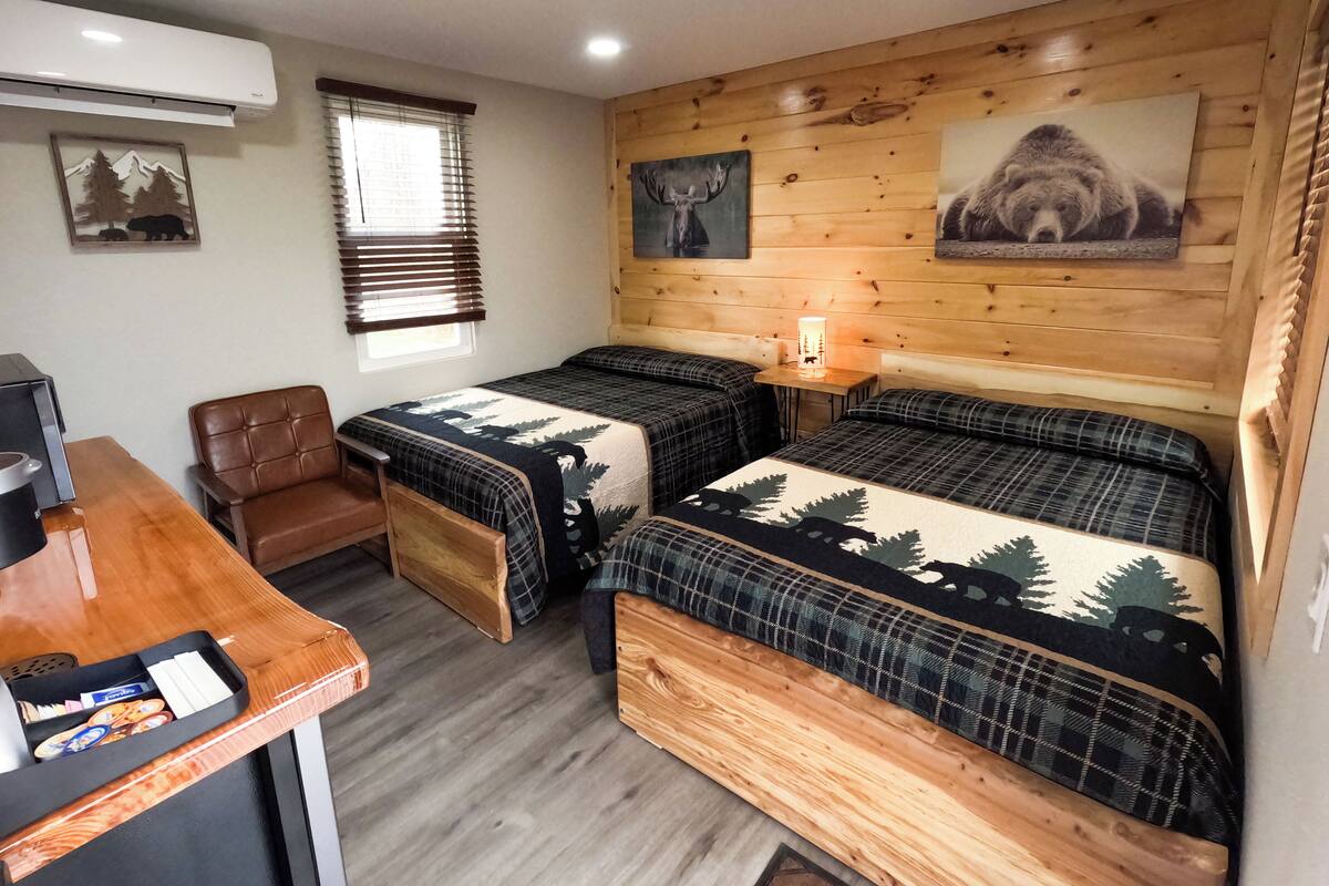 A cozy room with two beds, a desk, and a chair. There is a window with blinds, a wall-mounted air conditioner, and a lamp on a wooden table.
