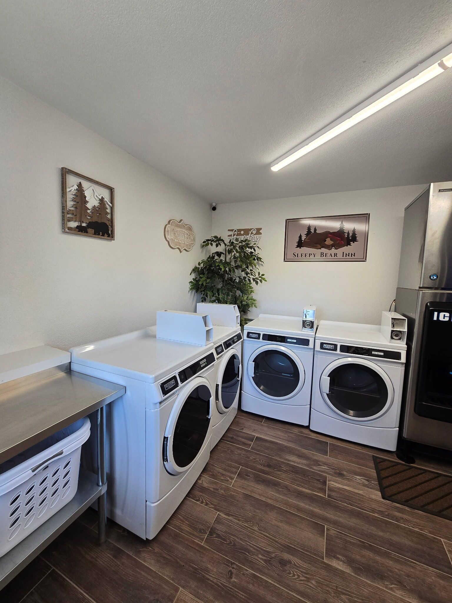 A laundry room with two front-loading washing machines, a dryer, and a refrigerator. There are wall decorations including a bear and a mountain scene.
